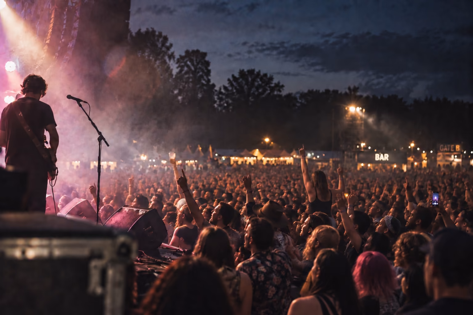 A festival crowd at dusk seen from the side of the stage, a guitarist in the foreground