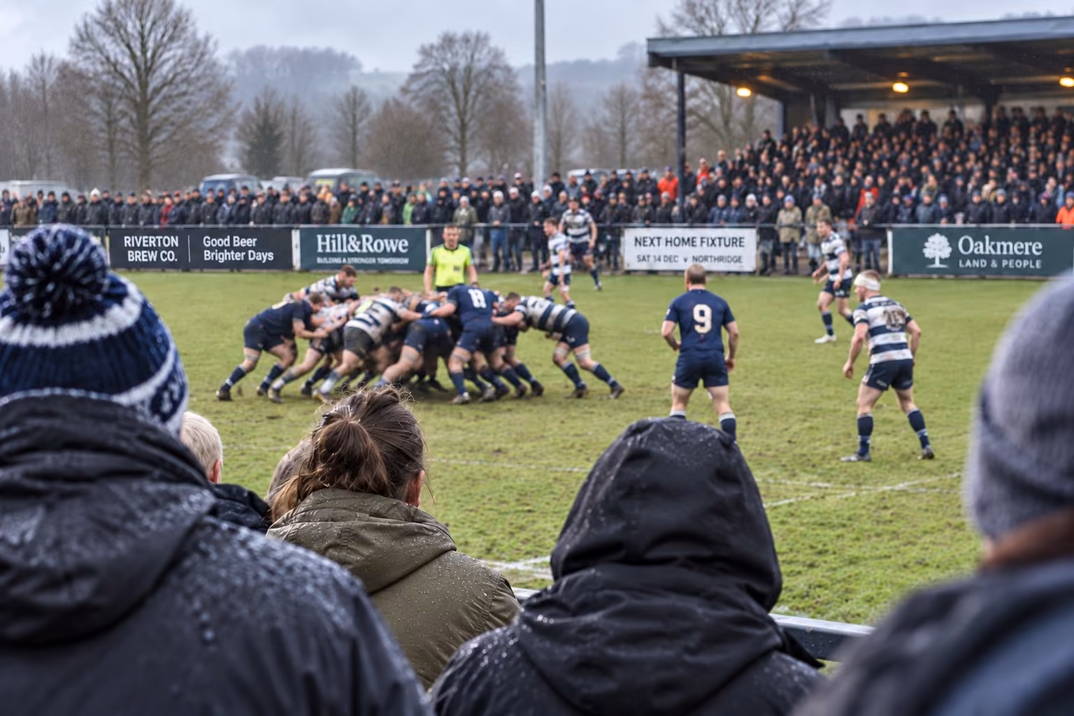 A club rugby scrum on a wet afternoon, seen over the shoulders of supporters at the rail